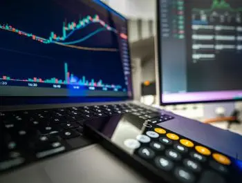 Financial analysis setup with laptop and secondary monitor showing stock charts, candlestick patterns, and a calculator,