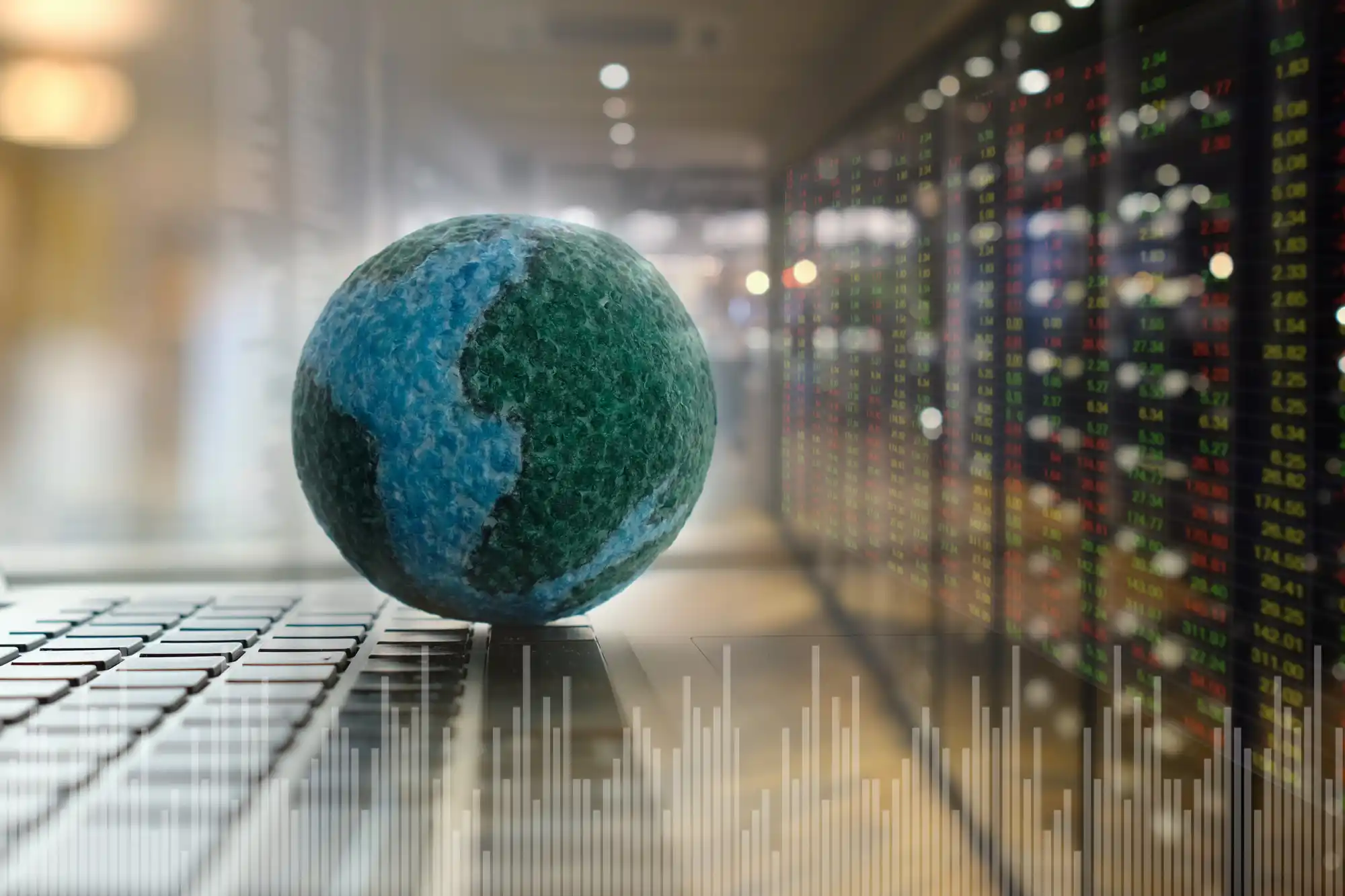 Small textured globe on a laptop keyboard with blurred financial data screens in the background, overlaid with graphs, representing global finance and digital trading.